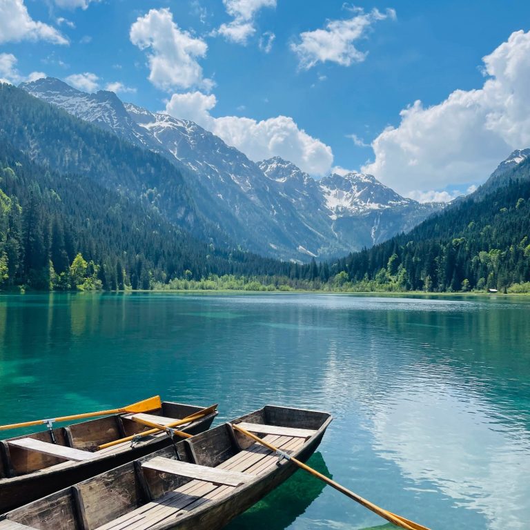 Türkisglitzernder Bergsee mit Bergkulisse, blauem Himmel, wenige weiße Wolken. Unten links zwei Holzruderboote mit Paddeln.