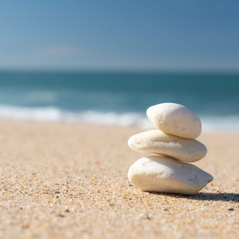 Drei helle Steine übereinander gestabelt an einem feinen Sandstrand. Im Hintergrund ist das flache Meer und ein blaue Himmel.